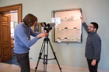one man looking through a camera on a tripod and the other holding up light to a cabinet